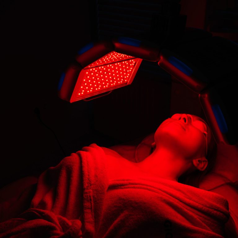 Relaxed female client having red LED light facial photodynamic therapy treatment in beauty salon. Young woman in protective glasses having non-invasive type of phototherapy. Beauty, wellness concept.